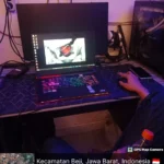 Teknisi mengetes performa laptop gaming dengan pencahayaan RGB di meja servis, disertai monitor eksternal dan casing PC di sampingnya. Kabel, colokan, dan peralatan kerja terlihat jelas di area belakang. Foto diambil malam hari di wilayah Beji, Depok