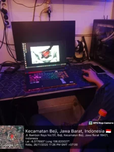 Teknisi mengetes performa laptop gaming dengan pencahayaan RGB di meja servis, disertai monitor eksternal dan casing PC di sampingnya. Kabel, colokan, dan peralatan kerja terlihat jelas di area belakang. Foto diambil malam hari di wilayah Beji, Depok