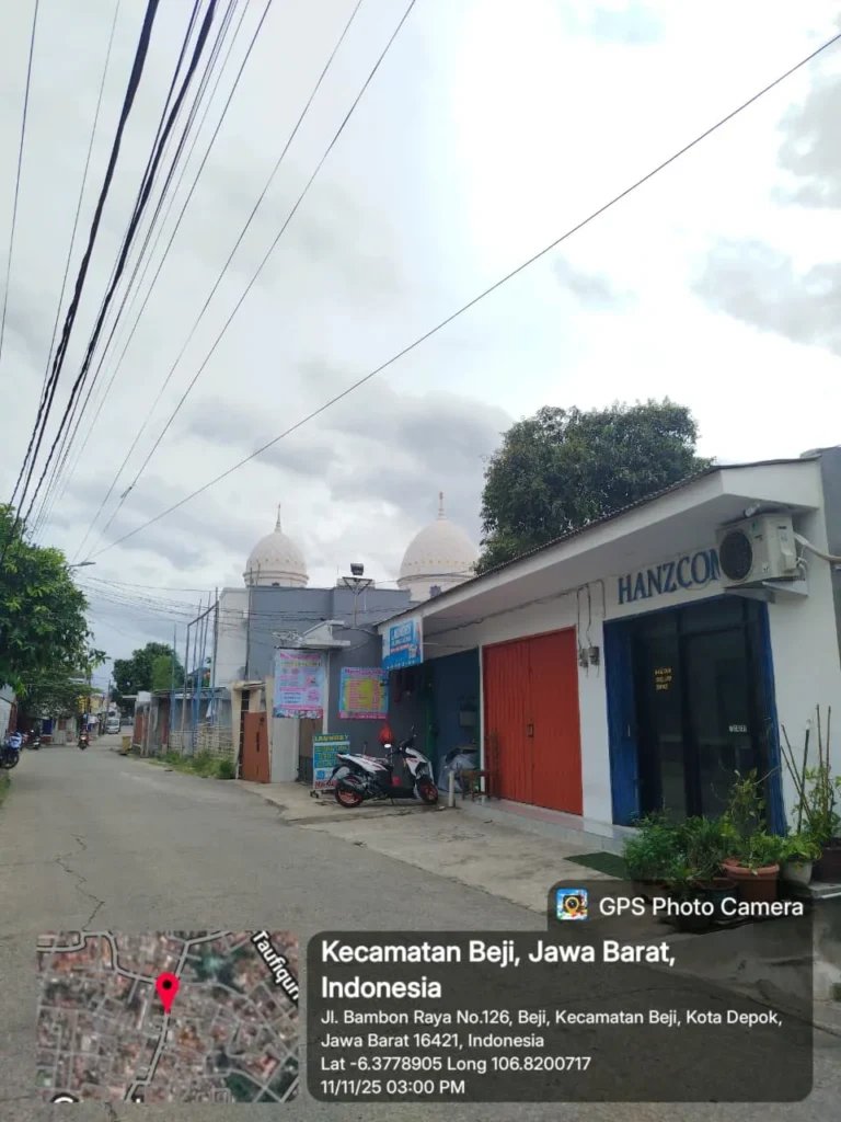 Foto suasana jalan Bambon Raya di Beji, Depok, menampilkan bangunan Hanz Com Laptop di sisi kanan jalan dengan pintu berwarna merah dan papan nama biru. Terlihat dua motor terparkir di depan toko, pohon besar di samping bangunan, serta menara masjid di latar belakang. Langit berawan dan lingkungan terlihat tenang seperti kawasan permukiman.”