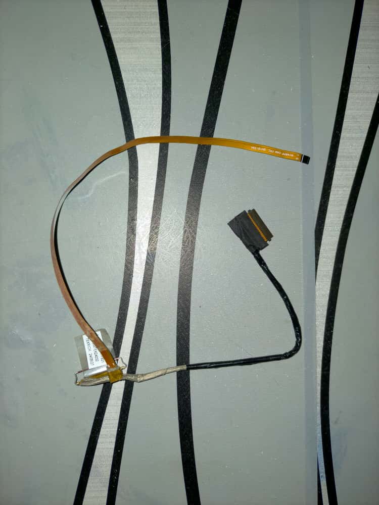 Kabel fleksibel layar laptop yang sudah aus dan retak pada bagian jalur, diperlihatkan di meja servis di Beji, Depok, sebagai contoh gejala tampilan berkedip atau layar gelap saat layar digerakkan