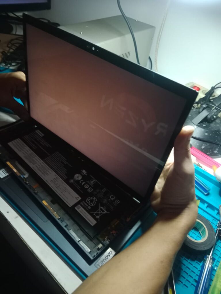 Proses pemeriksaan layar laptop yang sudah dilepas dari frame, memperlihatkan panel LCD dan bagian dalam perangkat dengan baterai serta jalur fleksibel. Teknisi sedang mengecek kondisi layar dan konektor untuk memastikan kerusakan pada tampilan