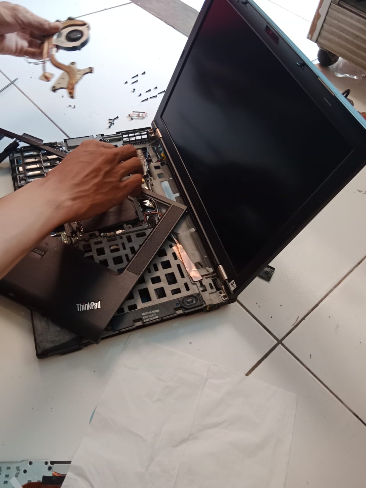 “Teknisi sedang membongkar laptop ThinkPad untuk membersihkan kipas dan sistem pendingin. Cover bawah sudah dilepas, komponen pendingin diangkat, dan layar terbuka lebar