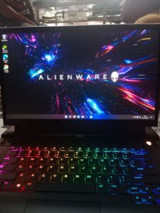 “Sebuah laptop gaming Alienware dengan layar menampilkan wallpaper efek cahaya berwarna neon dan keyboard RGB penuh, tampak dari sudut depan di atas meja kerja. Desain bodi tebal dan ventilasi besar khas Alienware terlihat jelas, menegaskan identitasnya sebagai laptop gaming kelas premium dengan sistem pendinginan kuat.”