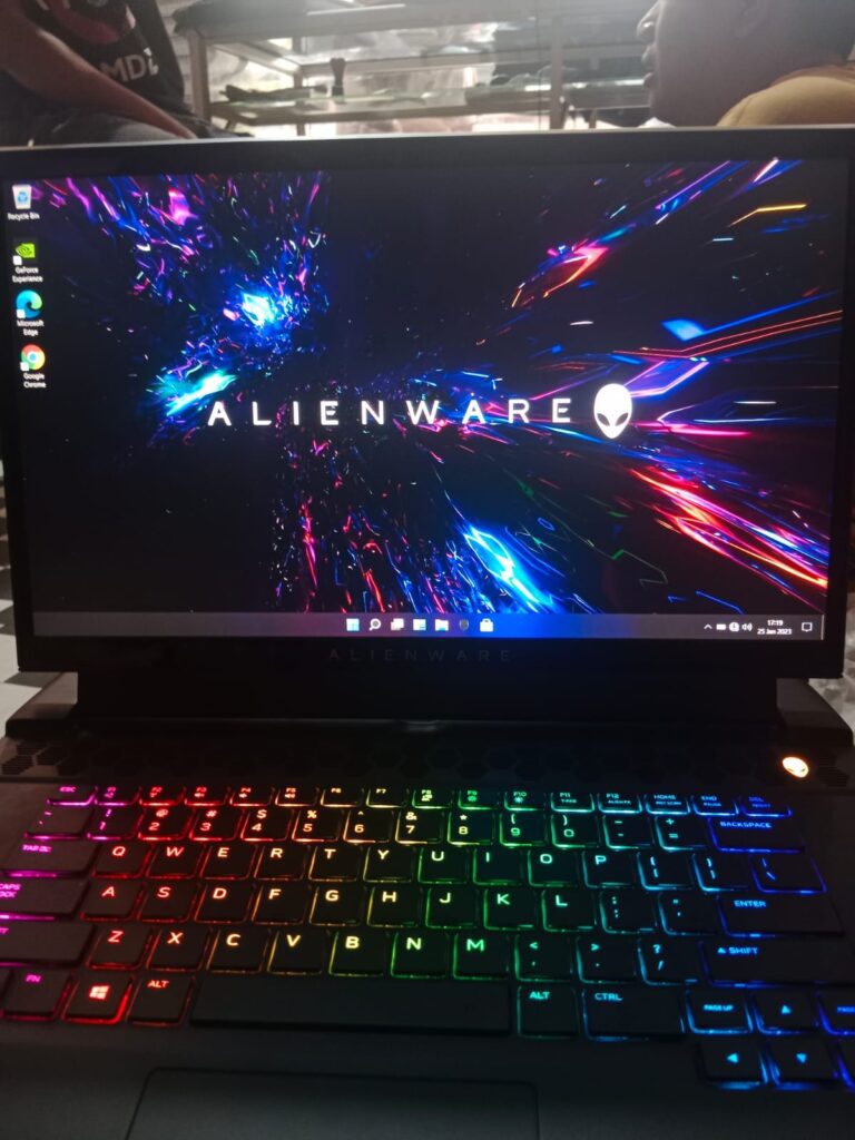 “Sebuah laptop gaming Alienware dengan layar menampilkan wallpaper efek cahaya berwarna neon dan keyboard RGB penuh, tampak dari sudut depan di atas meja kerja. Desain bodi tebal dan ventilasi besar khas Alienware terlihat jelas, menegaskan identitasnya sebagai laptop gaming kelas premium dengan sistem pendinginan kuat.”