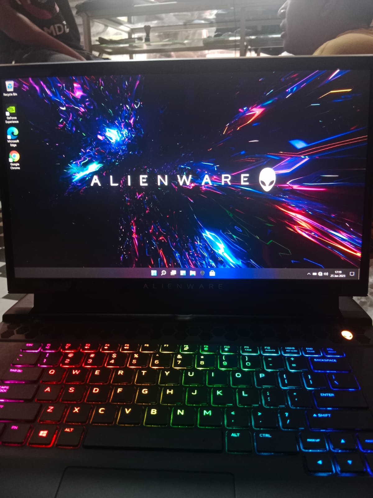 “Sebuah laptop gaming Alienware dengan layar menampilkan wallpaper efek cahaya berwarna neon dan keyboard RGB penuh, tampak dari sudut depan di atas meja kerja. Desain bodi tebal dan ventilasi besar khas Alienware terlihat jelas, menegaskan identitasnya sebagai laptop gaming kelas premium dengan sistem pendinginan kuat.”