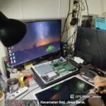 “Laptop dengan motherboard terbuka sedang diperiksa komponen VRM dan konektor layar; panel LCD dilepas, obeng presisi terlihat di tangan teknisi, dengan monitor eksternal dan alat solder di meja kerja.”