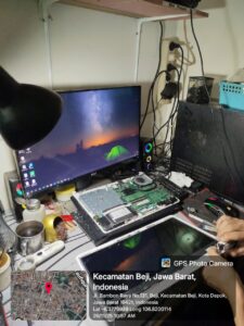 “Laptop dengan motherboard terbuka sedang diperiksa komponen VRM dan konektor layar; panel LCD dilepas, obeng presisi terlihat di tangan teknisi, dengan monitor eksternal dan alat solder di meja kerja.”