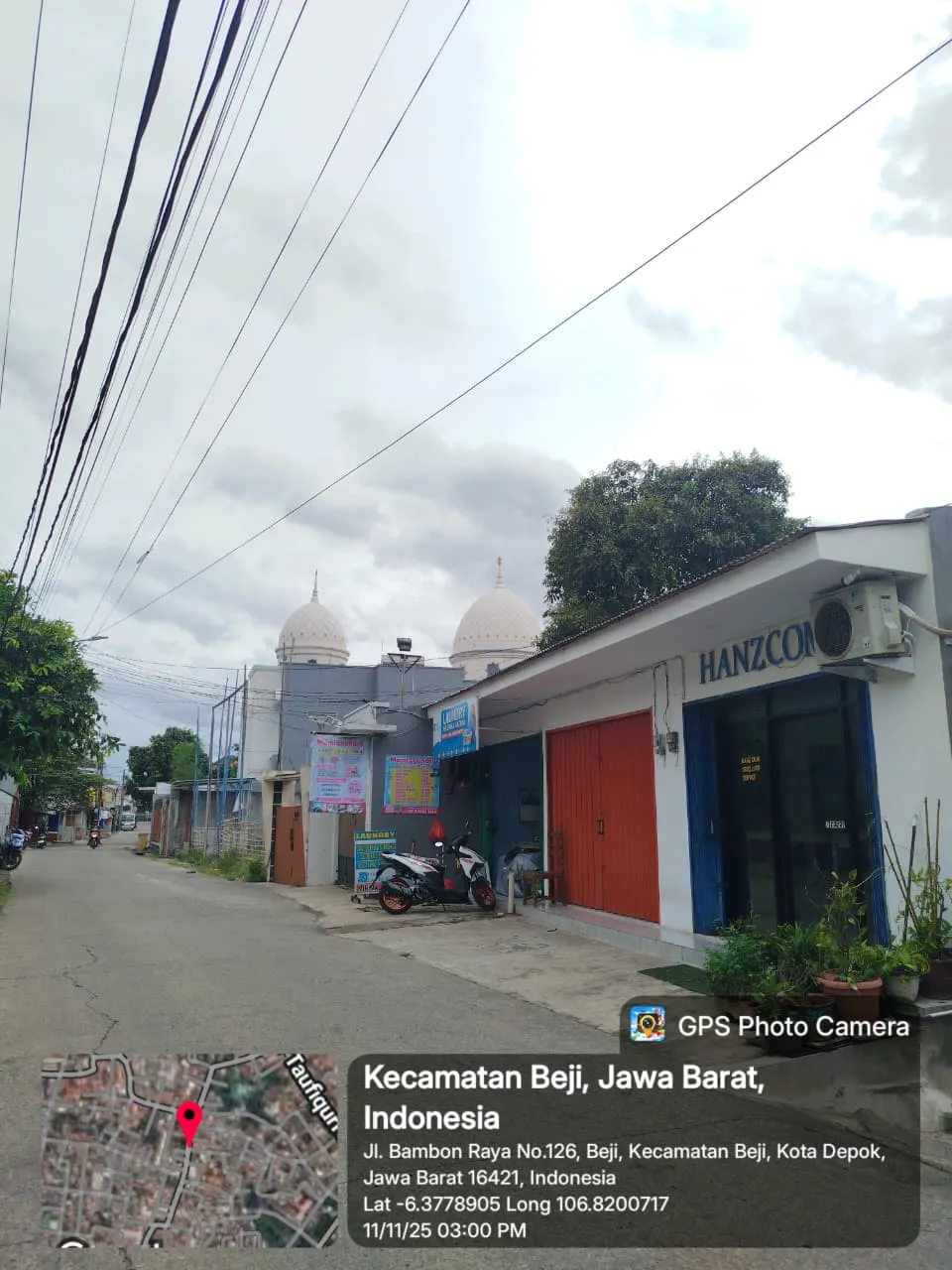 Foto suasana jalan Bambon Raya di Beji, Depok, menampilkan bangunan Hanz Com Laptop di sisi kanan jalan dengan pintu berwarna merah dan papan nama biru. Terlihat dua motor terparkir di depan toko, pohon besar di samping bangunan, serta menara masjid di latar belakang. Langit berawan dan lingkungan terlihat tenang seperti kawasan permukiman.”